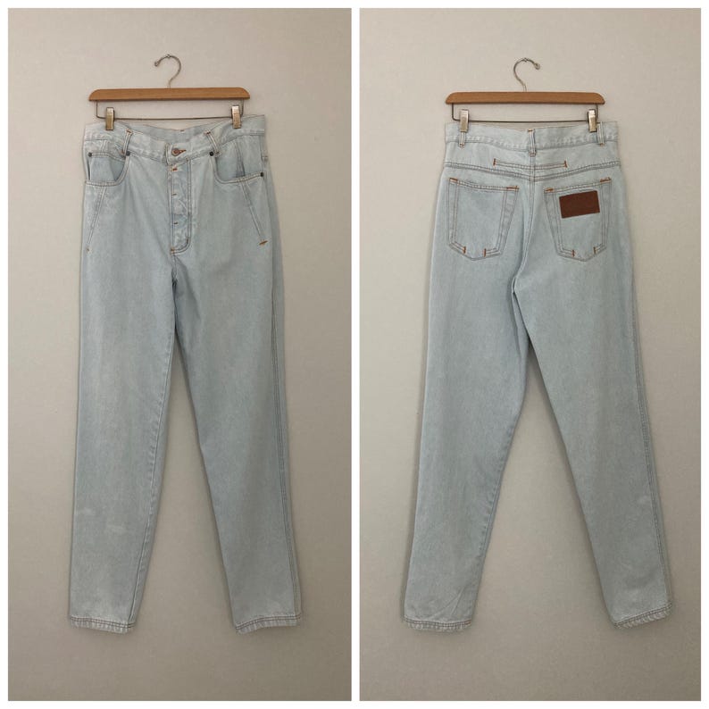 May include: Light blue denim jeans with a high waist and straight leg. The jeans have a button closure, pockets, and a brown leather patch on the back. The jeans are hanging on a wooden hanger.