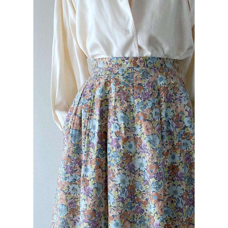 Puede incluir: Falda con estampado floral en colores pastel, con flores azules, moradas y melocot&oacute;n. La falda se combina con una camisa crema abotonada. La falda tiene un dise&ntilde;o plisado.