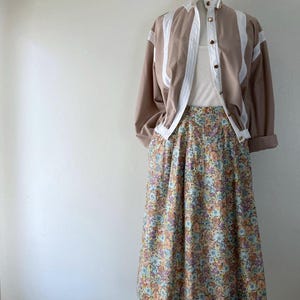 Puede incluir: Un atuendo vintage en un maniqu&iacute;. El conjunto incluye una chaqueta color canela con detalles blancos y botones dorados, una camiseta sin mangas blanca y una falda midi con estampado floral. La falda presenta un patr&oacute;n de peque&ntilde;as flores de varios colores.