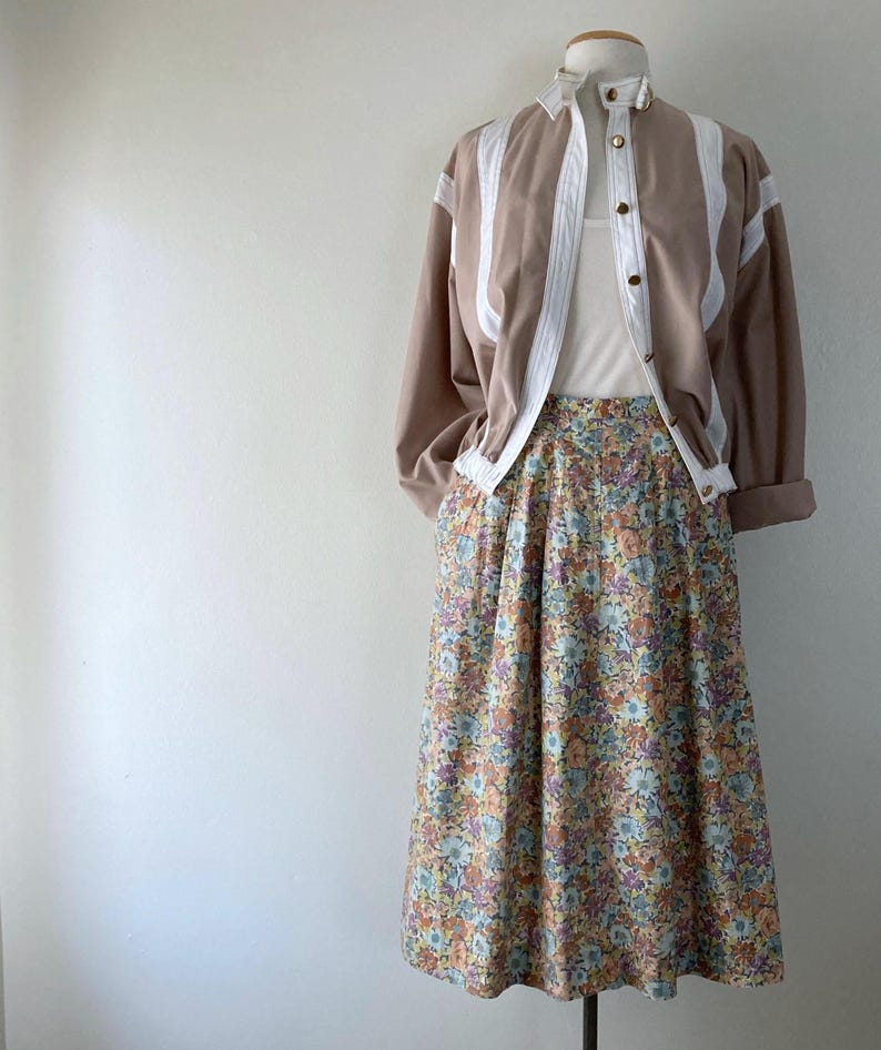Puede incluir: Un atuendo vintage en un maniqu&iacute;. El conjunto incluye una chaqueta color canela con detalles blancos y botones dorados, una camiseta sin mangas blanca y una falda midi con estampado floral. La falda presenta un patr&oacute;n de peque&ntilde;as flores de varios colores.