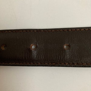May include: Close-up of a dark brown leather belt with visible stitching along the edges. The belt has several evenly spaced holes and a textured surface, suggesting a classic design.