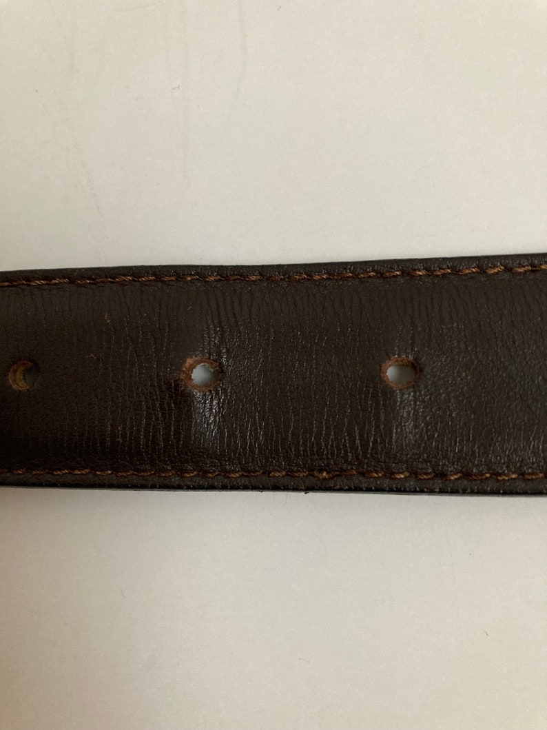 May include: Close-up of a dark brown leather belt with visible stitching along the edges. The belt has several evenly spaced holes and a textured surface, suggesting a classic design.