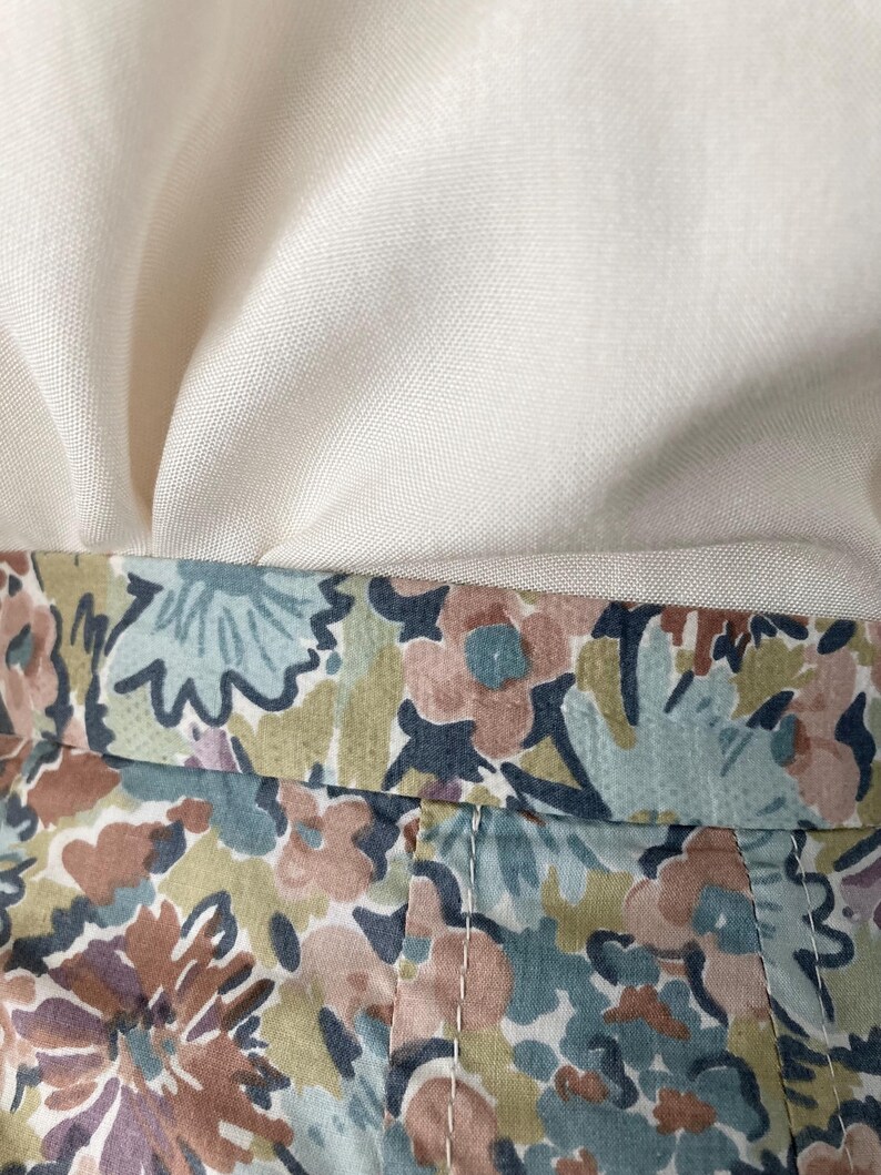 Puede incluir: Primer plano de tela con estampado floral en tonos azules, marrones y rosas. La tela est&aacute; cosida con hilo blanco. Encima de la tela floral hay una tela de color crema.