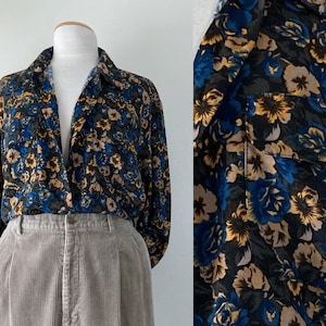 May include: A floral print blouse with a dark background, featuring blue, tan, and yellow flowers. The blouse has a collared neckline and long sleeves. Paired with light brown corduroy pants.