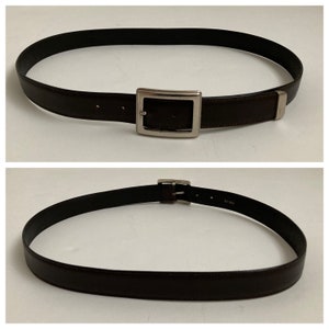 May include: A dark brown belt with a silver rectangular buckle. The belt is made of a smooth material and has a simple, classic design. The belt is shown in two views, one with the buckle centered and the other showing the belt's underside.
