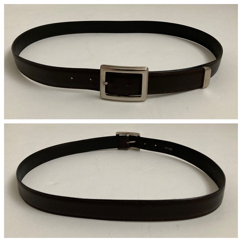 May include: A dark brown belt with a silver rectangular buckle. The belt is made of a smooth material and has a simple, classic design. The belt is shown in two views, one with the buckle centered and the other showing the belt's underside.