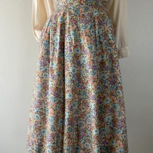 Puede incluir: Falda vintage con estampado floral en tonos pastel de azul, melocot&oacute;n, morado y amarillo. La falda se combina con una blusa crema de manga larga. La falda tiene una cintura alta y una silueta amplia plisada, creando un look retro.