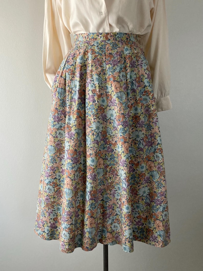Puede incluir: Falda vintage con estampado floral en tonos pastel de azul, melocot&oacute;n, morado y amarillo. La falda se combina con una blusa crema de manga larga. La falda tiene una cintura alta y una silueta amplia plisada, creando un look retro.