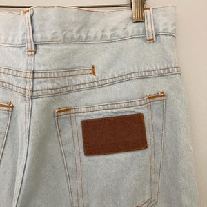 May include: Light blue denim jeans with a brown rectangular patch on the back pocket. The jeans have orange stitching and are hanging on a wooden hanger.