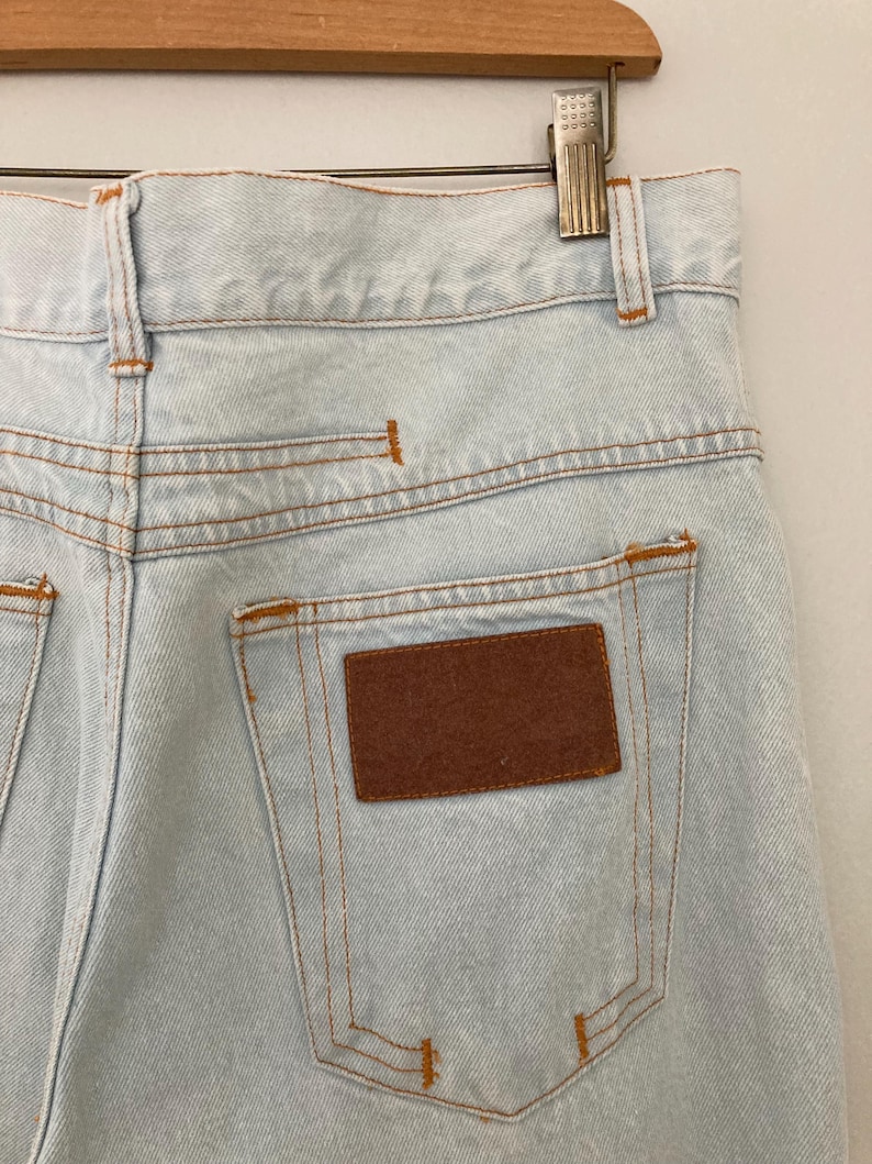 May include: Light blue denim jeans with a brown rectangular patch on the back pocket. The jeans have orange stitching and are hanging on a wooden hanger.