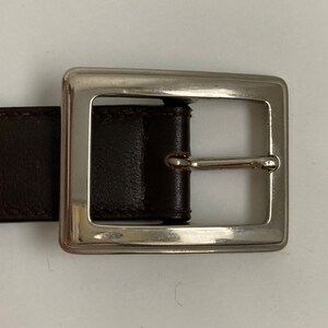May include: A close-up of a brown leather belt with a silver square buckle. The belt is made of dark brown leather and has a simple, classic design. The silver buckle is rectangular and has a shiny finish.