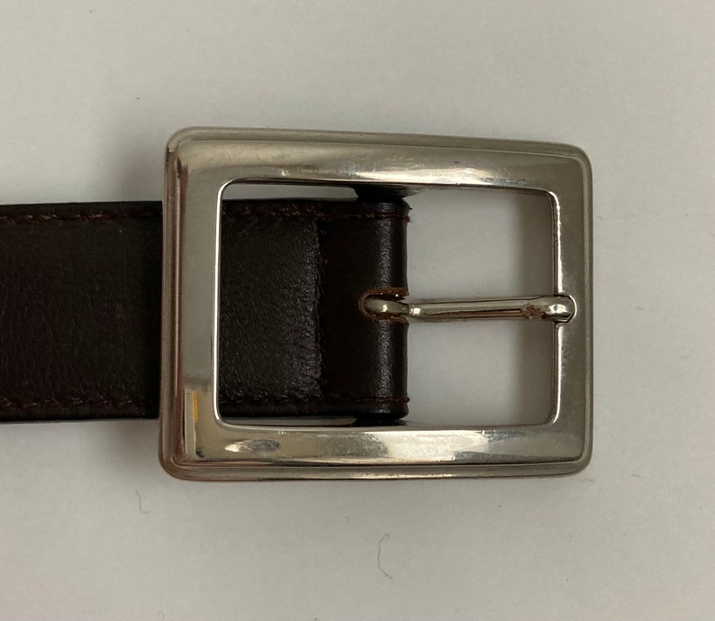 May include: A close-up of a brown leather belt with a silver square buckle. The belt is made of dark brown leather and has a simple, classic design. The silver buckle is rectangular and has a shiny finish.