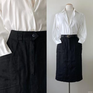 May include: A white collared shirt paired with a black knee-length skirt. The skirt features two front pockets and a button closure. A black belt with a gold buckle cinches the waist. The outfit is displayed on a mannequin.