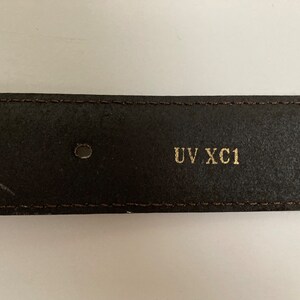 May include: Close-up of a dark brown leather belt with two holes and the text "UV XC1" in gold lettering. The belt has a stitched border and a slightly worn appearance.