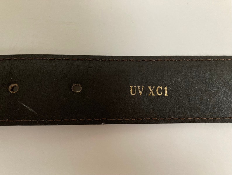 May include: Close-up of a dark brown leather belt with two holes and the text "UV XC1" in gold lettering. The belt has a stitched border and a slightly worn appearance.