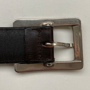 May include: A close-up of a brown leather belt with a silver square buckle. The buckle is engraved with "MCM MADE IN ITALY". A classic accessory, the belt is suitable for various outfits.