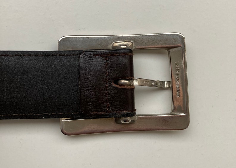 May include: A close-up of a brown leather belt with a silver square buckle. The buckle is engraved with "MCM MADE IN ITALY". A classic accessory, the belt is suitable for various outfits.