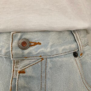 May include: Close-up of light blue denim jeans with a white t-shirt. The jeans have a button closure and orange stitching. The jeans have the text "BOYS RUGGLE" on the button and a small tag.