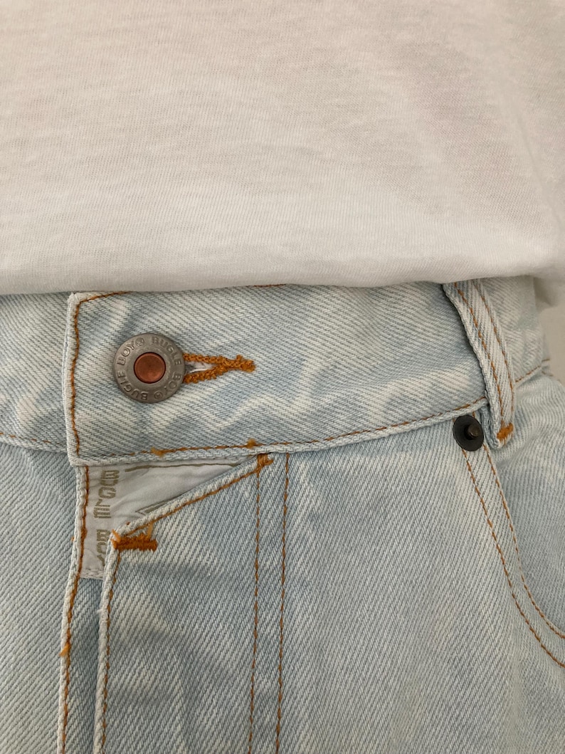May include: Close-up of light blue denim jeans with a white t-shirt. The jeans have a button closure and orange stitching. The jeans have the text "BOYS RUGGLE" on the button and a small tag.