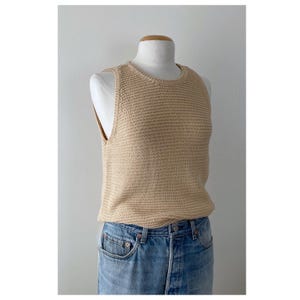 May include: A sleeveless beige knit tank top with a round neckline and a textured stitch pattern, paired with light blue denim jeans featuring a button fly closure and copper-colored rivets at the pockets and waistline.