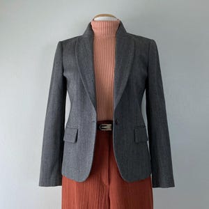 May include: A gray blazer with a subtle herringbone pattern, paired with a peach turtleneck and rust-colored pants. The blazer has a classic notched lapel and two front pockets. A brown belt cinches the waist.