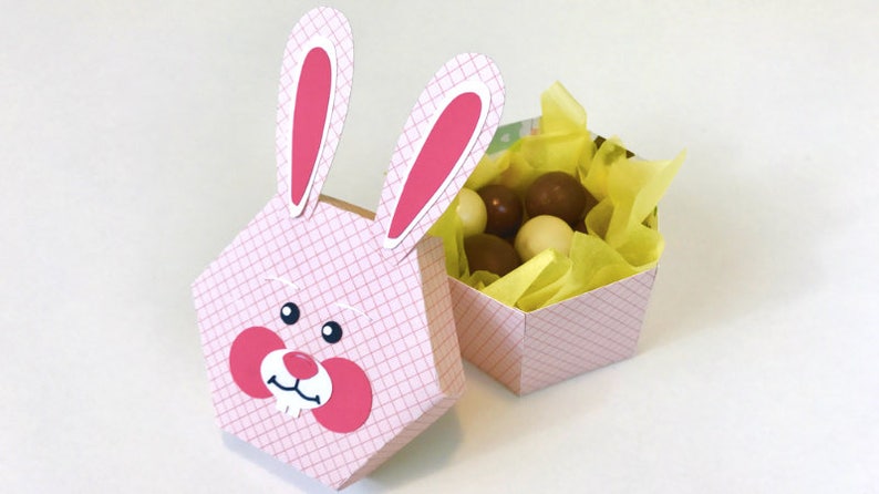 May include: A pink hexagonal box with a bunny face on the front. The box is open and filled with chocolate candies and yellow tissue paper.