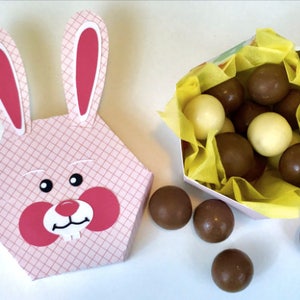 Puede incluir: Una caja con forma de conejo rosa y blanca con un patrón de cuadros rosa y blanco. La caja está llena de caramelos de chocolate, algunos son blancos y otros son marrones. Los caramelos se están derramando de la caja sobre una superficie blanca.