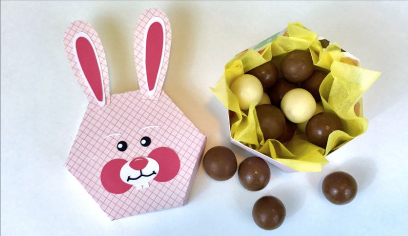 May include: A pink and white bunny-shaped box with a pink and white checkered pattern. The box is filled with chocolate candies, some are white and some are brown. The candies are spilling out of the box onto a white surface.