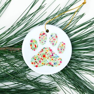 May include: White ceramic Christmas ornament shaped like a paw print, decorated with a colorful floral pattern.