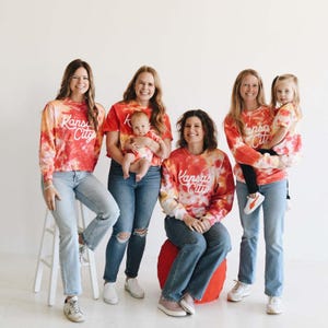 May include: Five people, including a young child, are wearing tie-dye shirts with the text "Kansas City" in white. The shirts are in shades of red, orange, and yellow. The people are standing in front of a white wall.