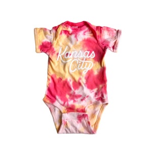 May include: A pink and yellow tie-dye baby onesie with the text "Kansas City" in white.