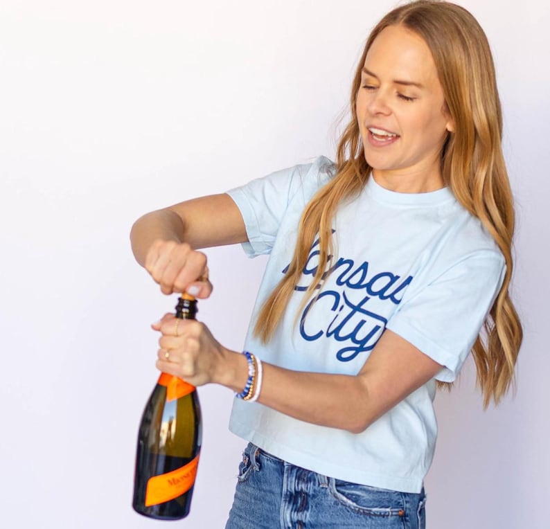 May include: A woman wearing a light blue t-shirt with "Kansas City" written in blue script on the front, is popping open a bottle of champagne. She is wearing blue jeans and a bracelet on her wrist.