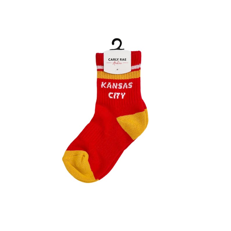 May include: Red and gold Kansas City socks with the text "KANSAS CITY" in white. The socks have a white and gold stripe at the top and a black tag with the brand name "Carly Rae Studio".