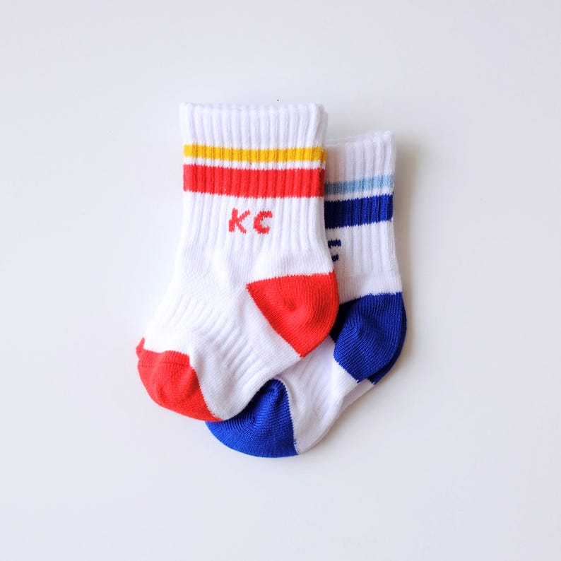 Peut inclure: Deux paires de chaussettes de sport blanches avec des rayures et des accents color&eacute;s. Une paire a des rayures rouges, jaunes et rouges avec "KC" en rouge. L'autre a des rayures et des accents bleus. Les chaussettes ont des orteils rouges et bleus.