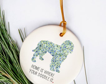 Könnte beinhalten: Weißer Keramik-Anhänger mit einer Doodle-Hundesilhouette aus blauen und grünen Blumen. Der Anhänger hat eine goldene Schnur zum Aufhängen und den Text "Home is where your doodle is" mit einem kleinen roten Herzen.