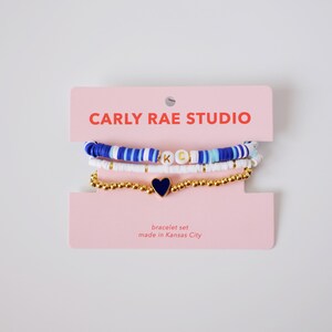 Könnte beinhalten: Armband-Set auf einer rosa Karte mit dem Text "CARLY RAE STUDIO". Die Armbänder umfassen blaue und weiße Scheibenperlen, goldene Perlen und eine goldene Kette mit einem marineblauen Herzanhänger. Die Karte sagt auch "Armband-Set".