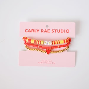 May include: A bracelet set featuring three bracelets. One bracelet has red and yellow beads with the letters "KC". Another bracelet is red, and the third is gold with a red heart charm. The packaging reads "CARLY RAE STUDIO".