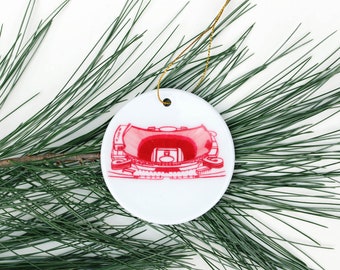 Op de afbeelding: Witte kerstbal van keramiek met een rode illustratie van een stadion.