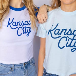 May include: Two women wearing t-shirts with the text "Kansas City" in blue script. The shirt on the left is a white and blue baseball style shirt. The shirt on the right is light blue.