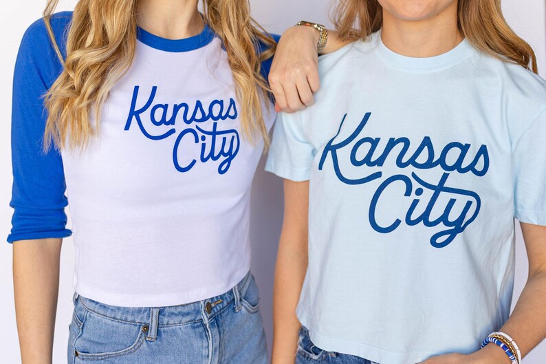 May include: Two women wearing t-shirts with the text "Kansas City" in blue script. The shirt on the left is a white and blue baseball style shirt. The shirt on the right is light blue.