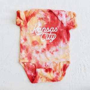 May include: A red and yellow tie-dye baby onesie with the text "Kansas City" in white.