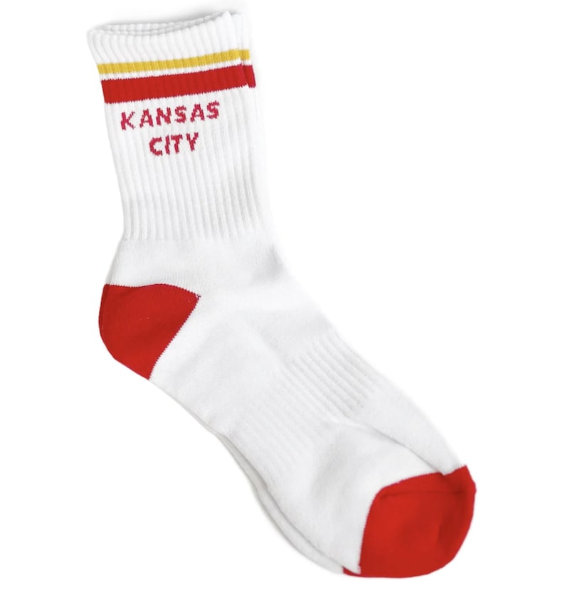 May include: White crew socks with red and yellow stripes at the top. The socks have a red heel and toe and say "Kansas City" in red letters.