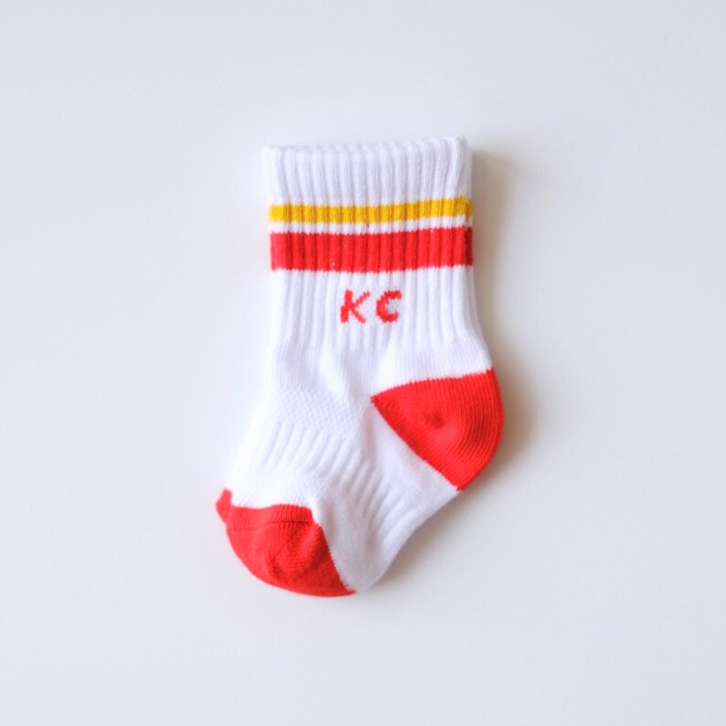 Peut inclure: Chaussette de b&eacute;b&eacute; blanche avec des rayures rouges et jaunes en haut et des accents rouges sur le bout et le talon. Les lettres "KC" sont brod&eacute;es en rouge sur le devant de la chaussette.
