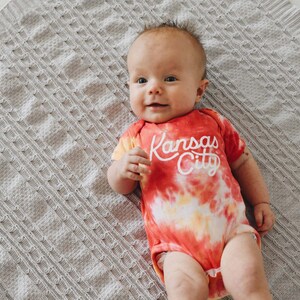 May include: A baby wearing a red, yellow and orange tie-dye onesie with the text "Kansas City" in white. The baby is lying on a grey blanket.