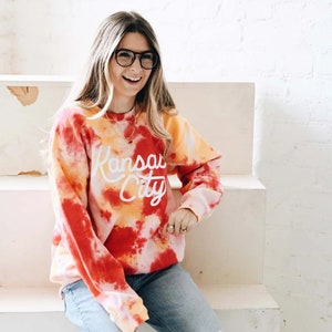 May include: A red, orange, and yellow tie-dye sweatshirt with the text "Kansas City" in white lettering. The sweatshirt is worn by a person with long blonde hair and glasses.