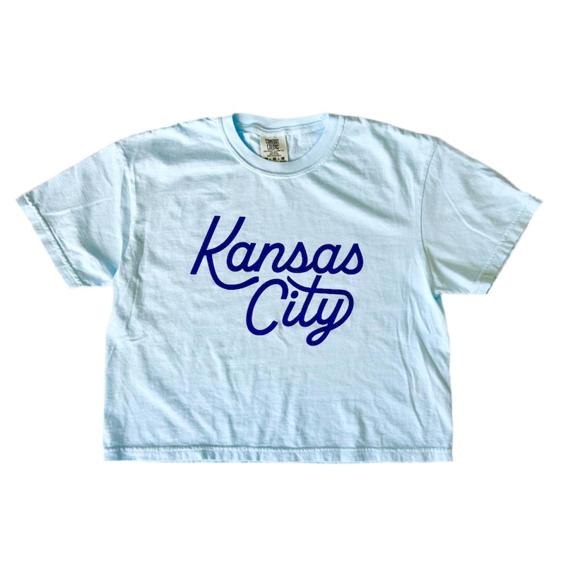 May include: A light blue cropped t-shirt with a navy blue "Kansas City" graphic in a retro script font.