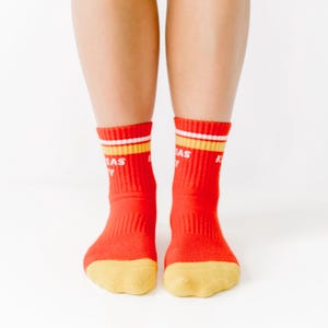 May include: A pair of red socks with yellow and white stripes at the top and yellow toes. The socks have the word "KANSAS" printed on them in white letters. The socks are worn by a person.