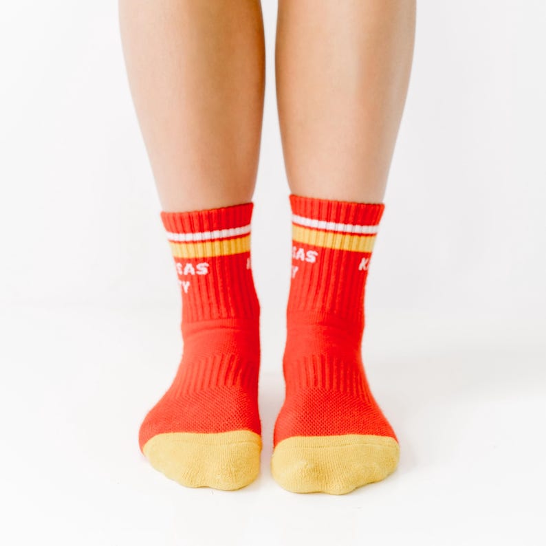 May include: A pair of red socks with yellow and white stripes at the top and yellow toes. The socks have the word "KANSAS" printed on them in white letters. The socks are worn by a person.
