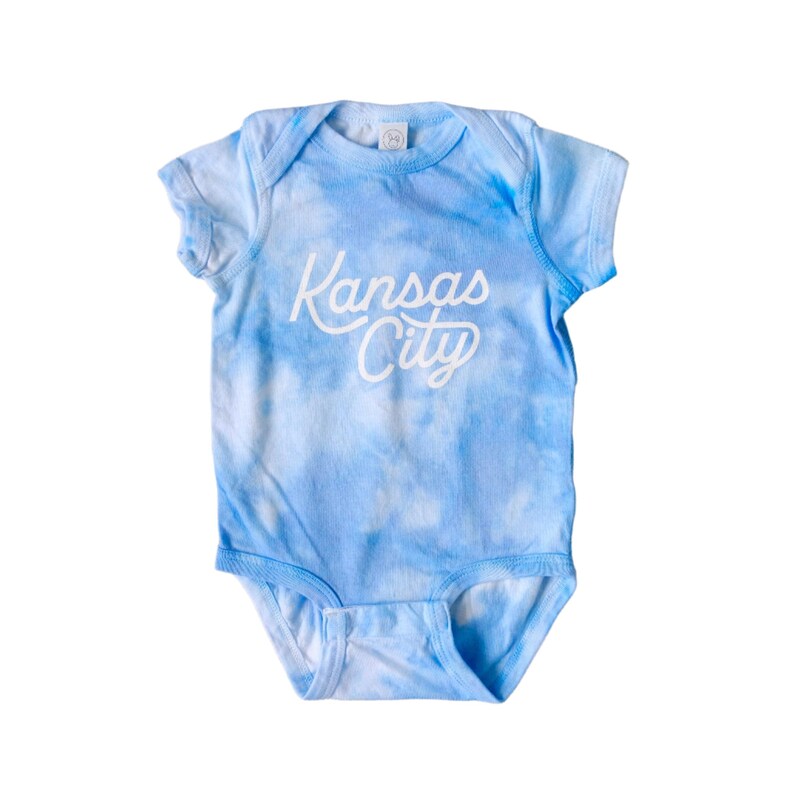 K&ouml;nnte beinhalten: Ein Baby-Body mit einem Batikmuster in Blau- und Wei&szlig;t&ouml;nen. Der Schriftzug "Kansas City" ist in wei&szlig;er Schrift aufgedruckt. Der Body hat kurze &Auml;rmel und einen Druckknopfverschluss.