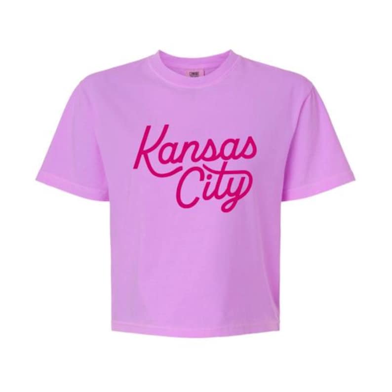 Puede incluir: Una camiseta de manga corta de color morado claro con el texto Kansas City impreso en una letra cursiva de color rosa oscuro en el centro del pecho. La prenda tiene un cuello redondo y un corte est&aacute;ndar con un dobladillo recto.
